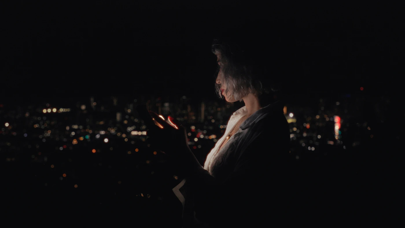 Joueuse éclairée par l'écran de son téléphone en soirée, silhouette de la ville en arrière-plan